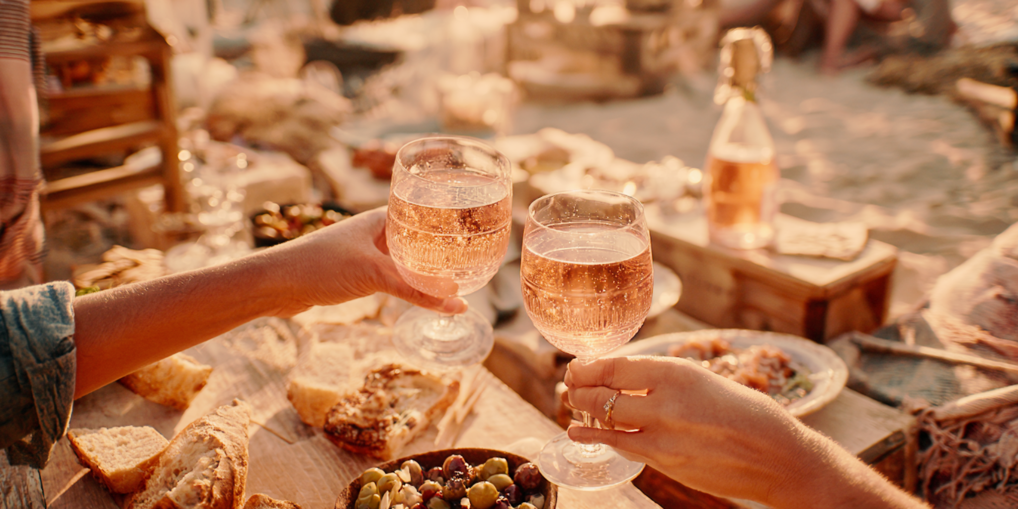 Two people toasting with glasses of rose wine at a beachside table with food.