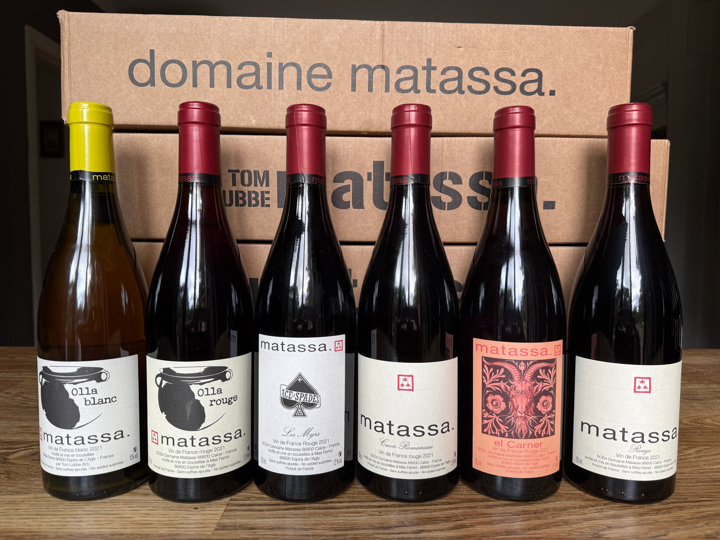 Six bottles of Domaine Matassa natural wine lined up on a wooden surface, with cardboard boxes behind them reading “domaine matassa.” Includes Olla Blanc, Olla Rouge, Ace of Spades, Romanissa, El Carner, and Rouge.