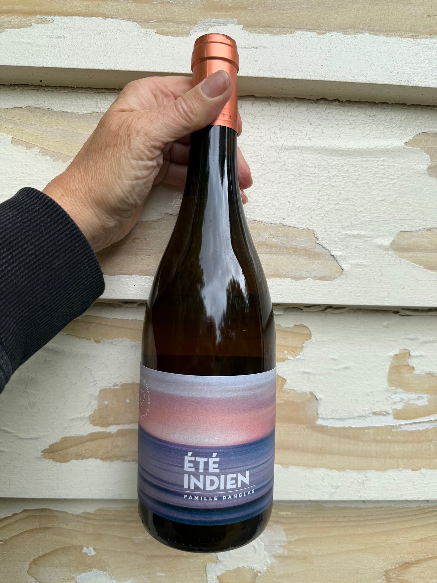 A hand holding a bottle of Château de Paraza Été Indien orange wine against a rustic cream and wood-textured wall, showcasing the wine’s elegant label and copper capsule.