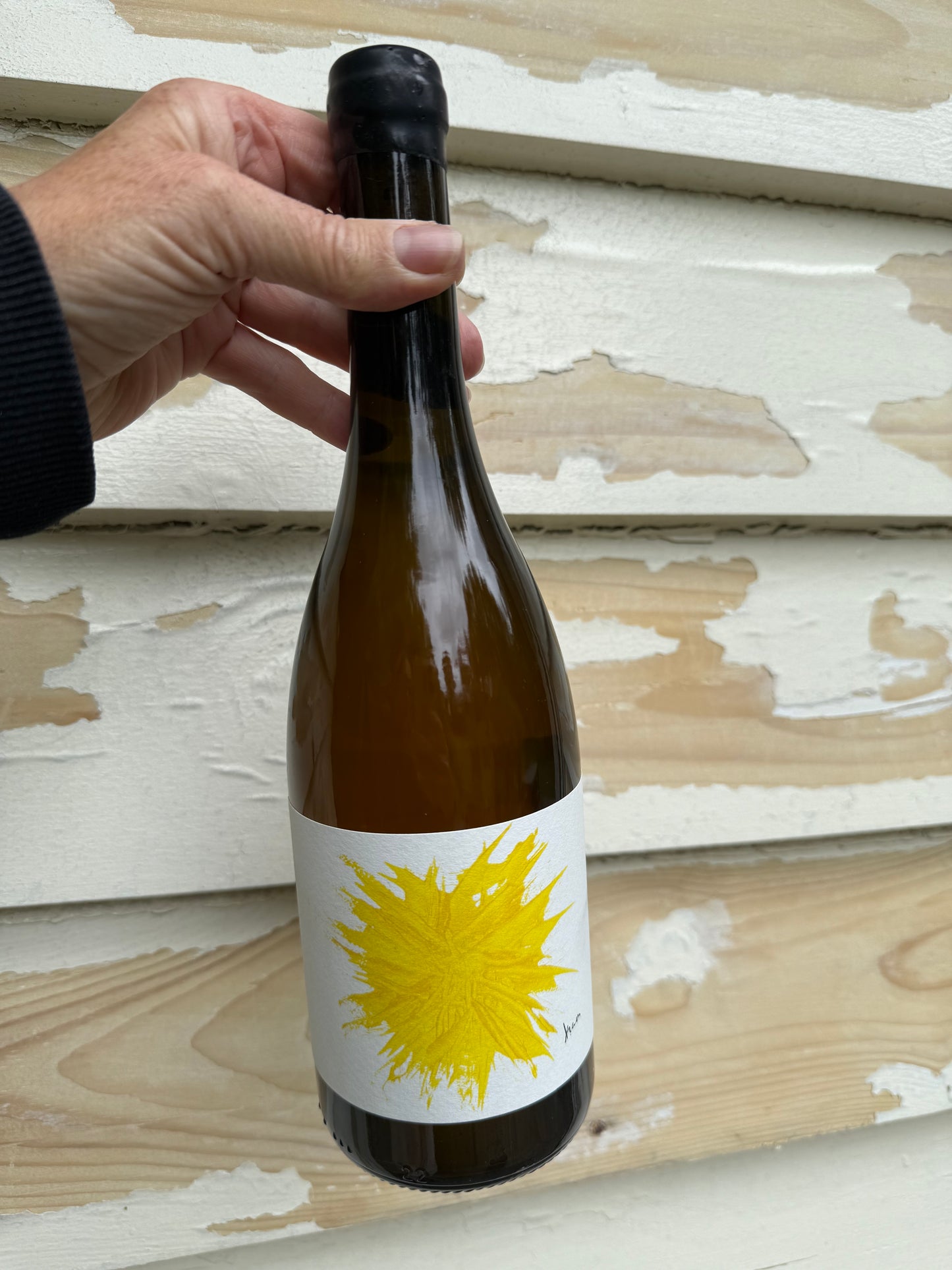 A hand holding a bottle of Gilles Troullier Maceration Orange wine with a striking yellow label, set against a distressed cream wooden backdrop. The bottle showcases the natural amber hue of the wine.