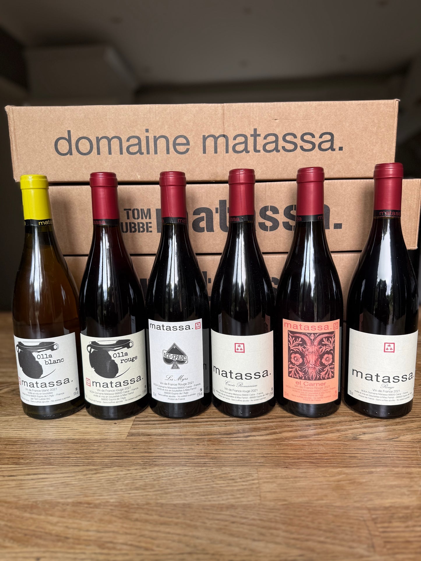 Six bottles of Domaine Matassa natural wine lined up on a wooden surface, with cardboard boxes behind them reading “domaine matassa.” Includes Olla Blanc, Olla Rouge, Ace of Spades, Romanissa, El Carner, and Rouge.