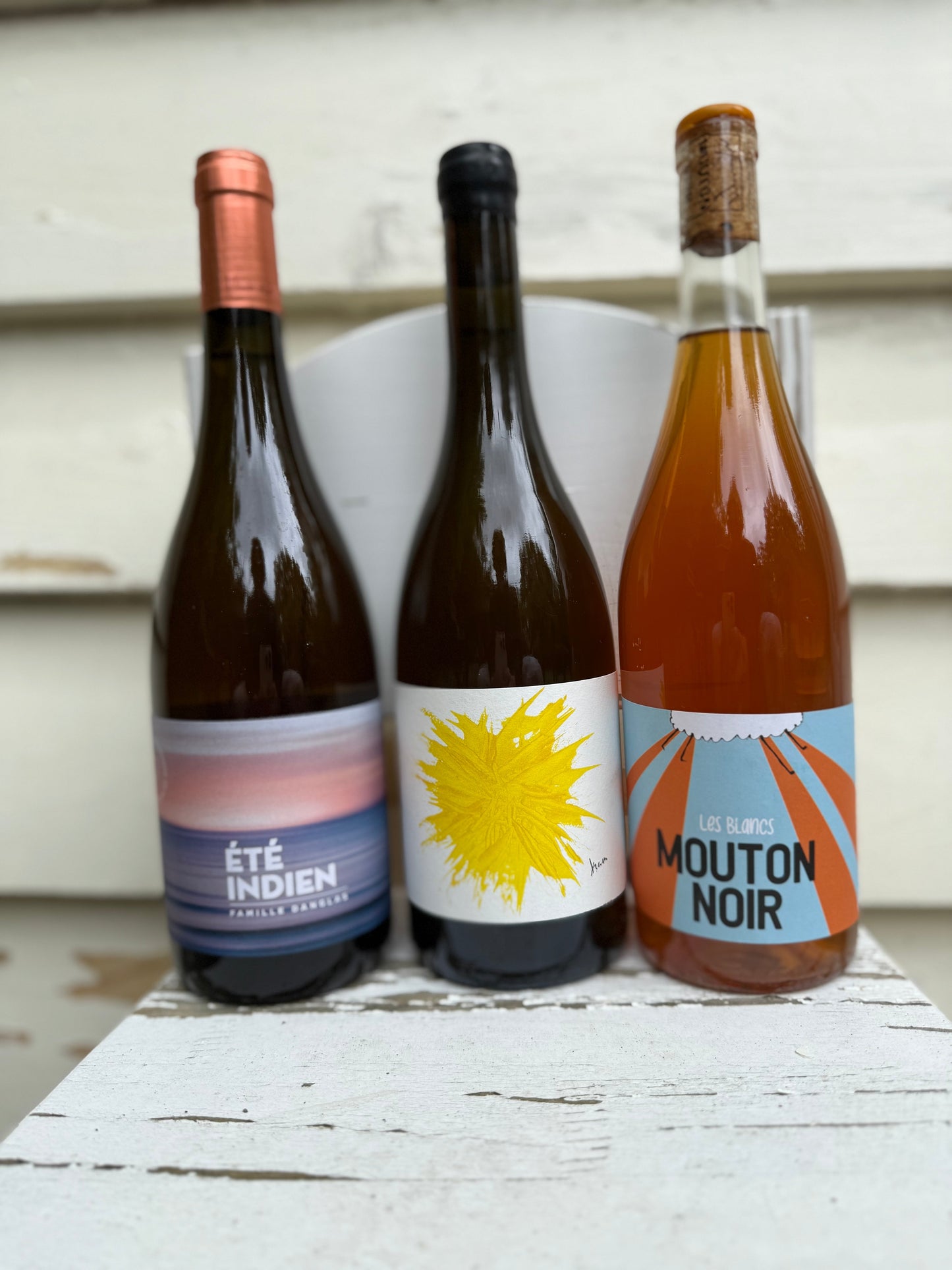 Three bottles of natural orange wine — Été Indien, Colline de l’Hirondelle Bleu, and Mouton Noir Les Blancs — displayed together as a curated gift set on a rustic white table.