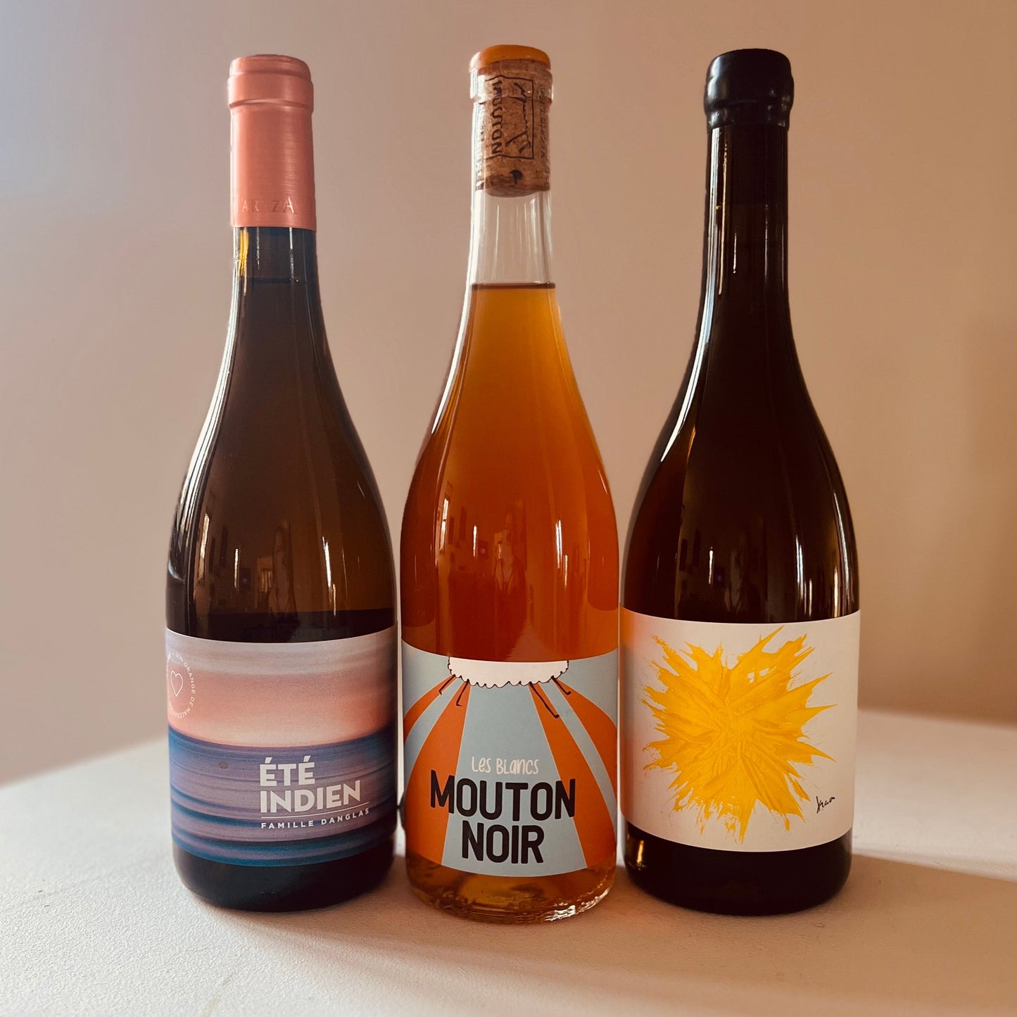 Three bottles of Taste Union orange wines—Été Indien, Les Blancs Mouton Noir, and a third sun-burst label—standing side by side on a white table against a softly lit neutral background.
