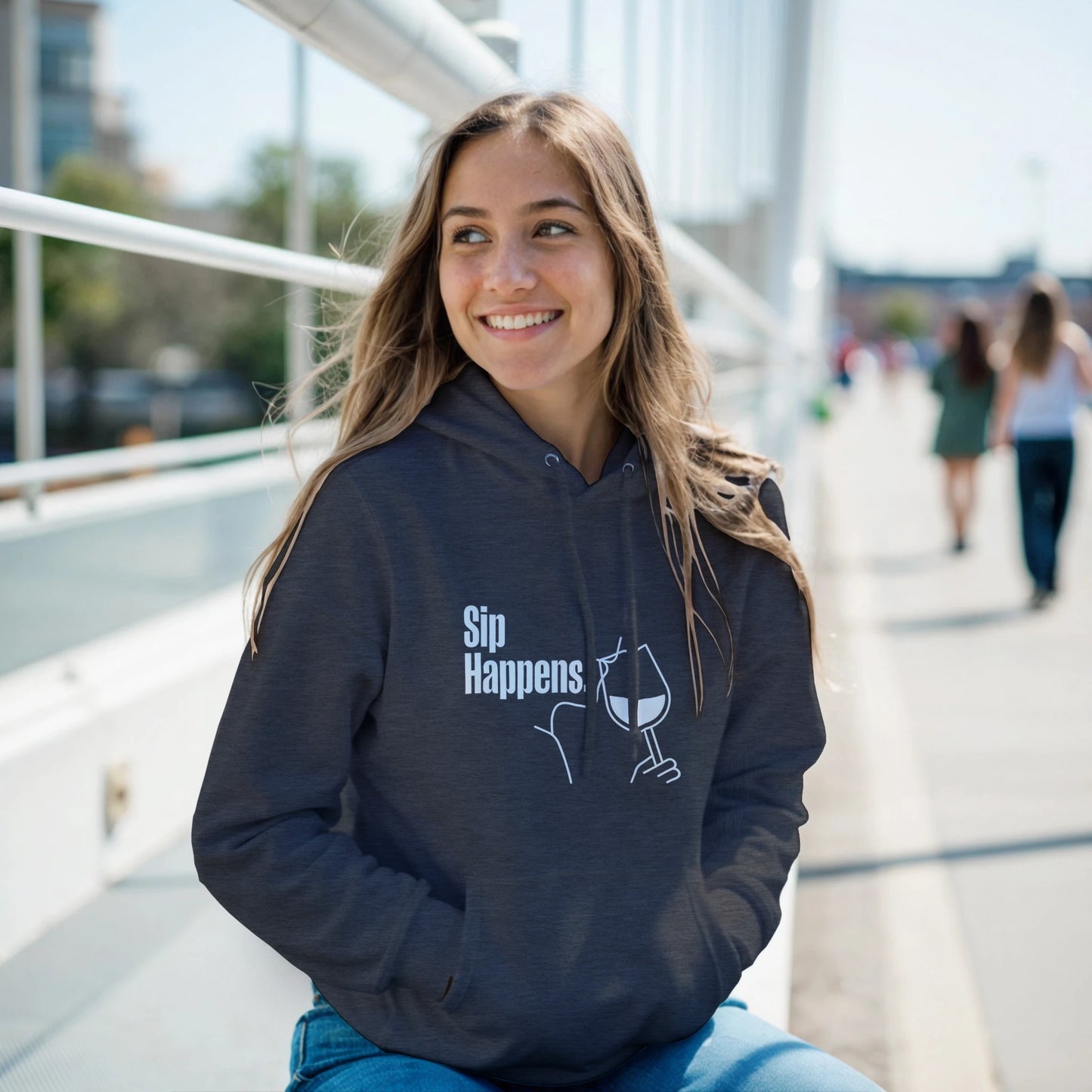 Woman wearing a grey hoodie with 'Sip Happens' text and graphic, sitting outdoors.