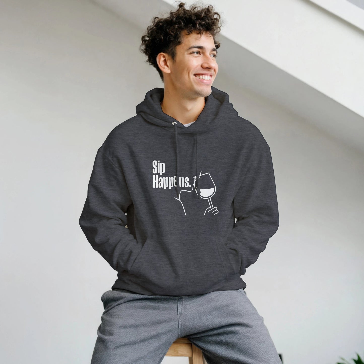 Man wearing a dark gray hoodie with text and graphics, sitting on a white surface.