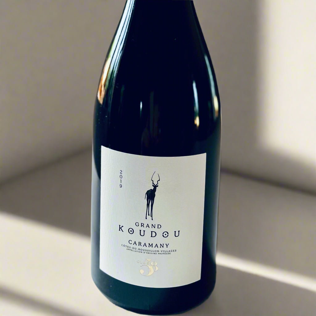 A close-up image of the Grand Koudou Caramany 2019 wine bottle featuring a clean white label with a black silhouette of a kudu antelope. The label highlights the wine’s name, vintage, and appellation. The bottle rests on a neutral-coloured background, emphasising its elegant design and minimalistic aesthetic.