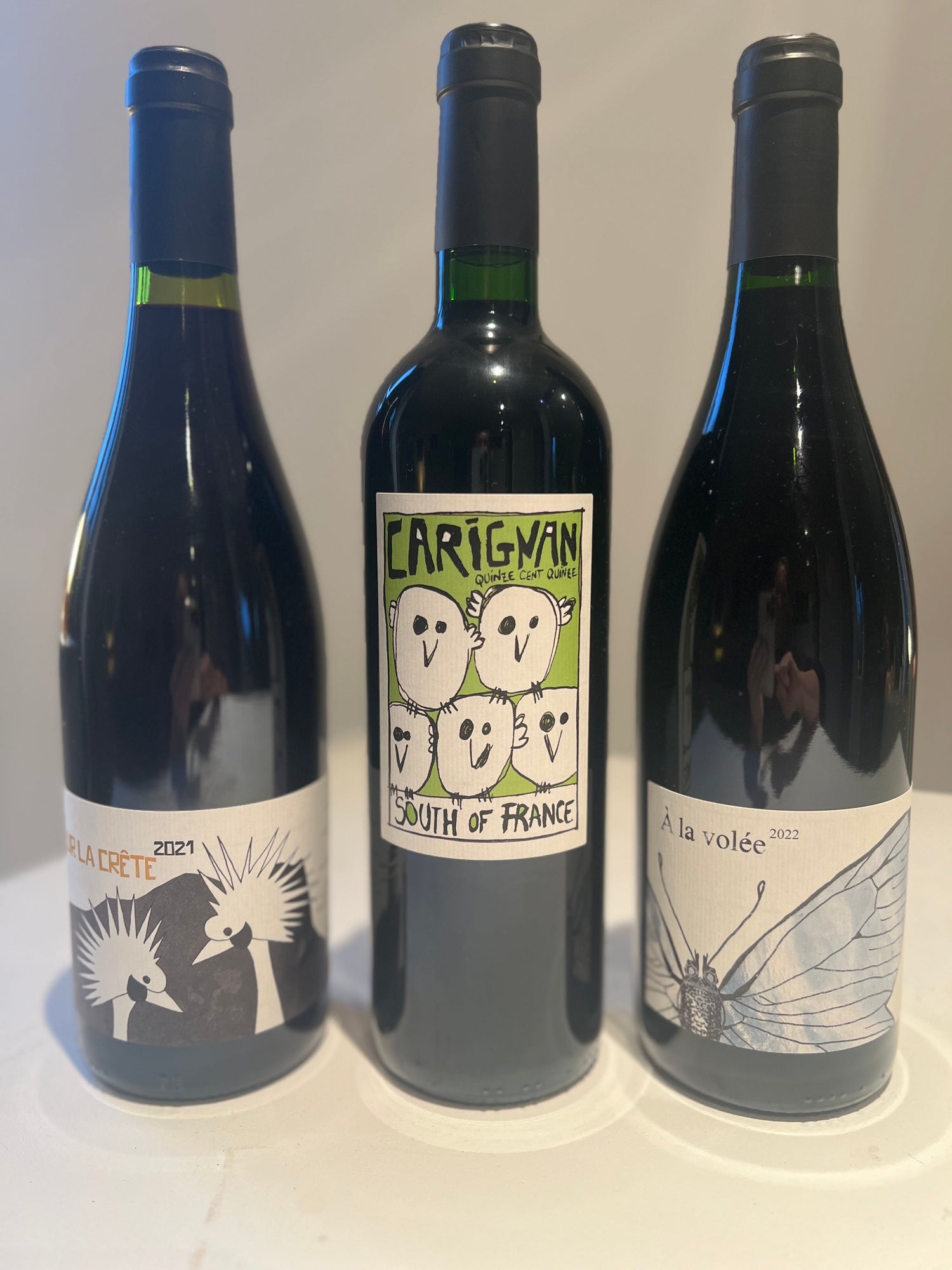 Three bottles of biodynamic red wine from Colline de l'Hirondelle: "Sur La Crête 2021" with birds, "Carignan | South of France" with smiling faces, and "A la volée 2022" with a dragonfly. Showcasing creativity, sustainability, and quality craftsmanship.