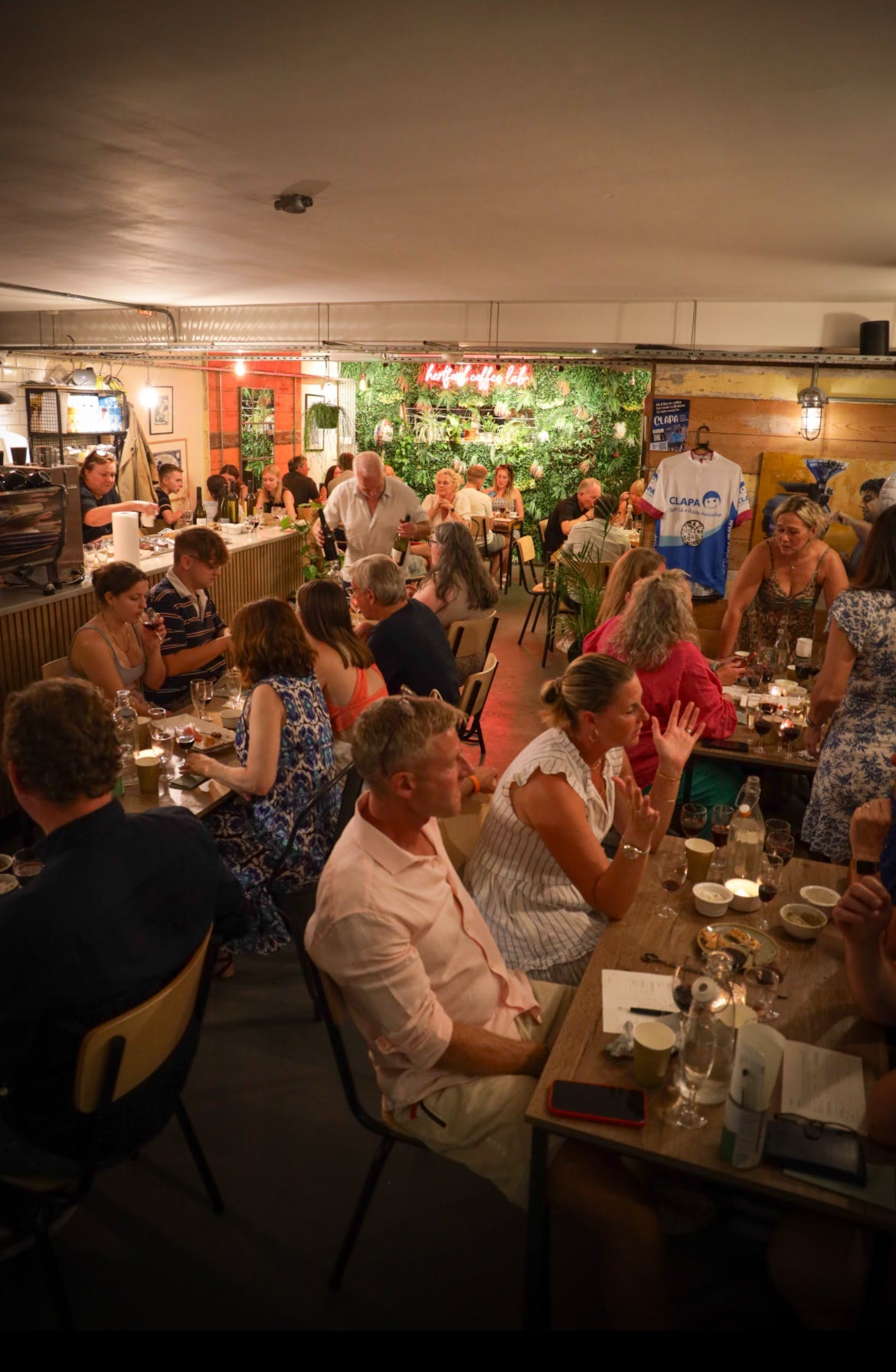 A lively wine tasting event at Hertford Coffee Lab, featuring guests seated at tables enjoying wine and food pairings in a warm, plant-filled ambiance.