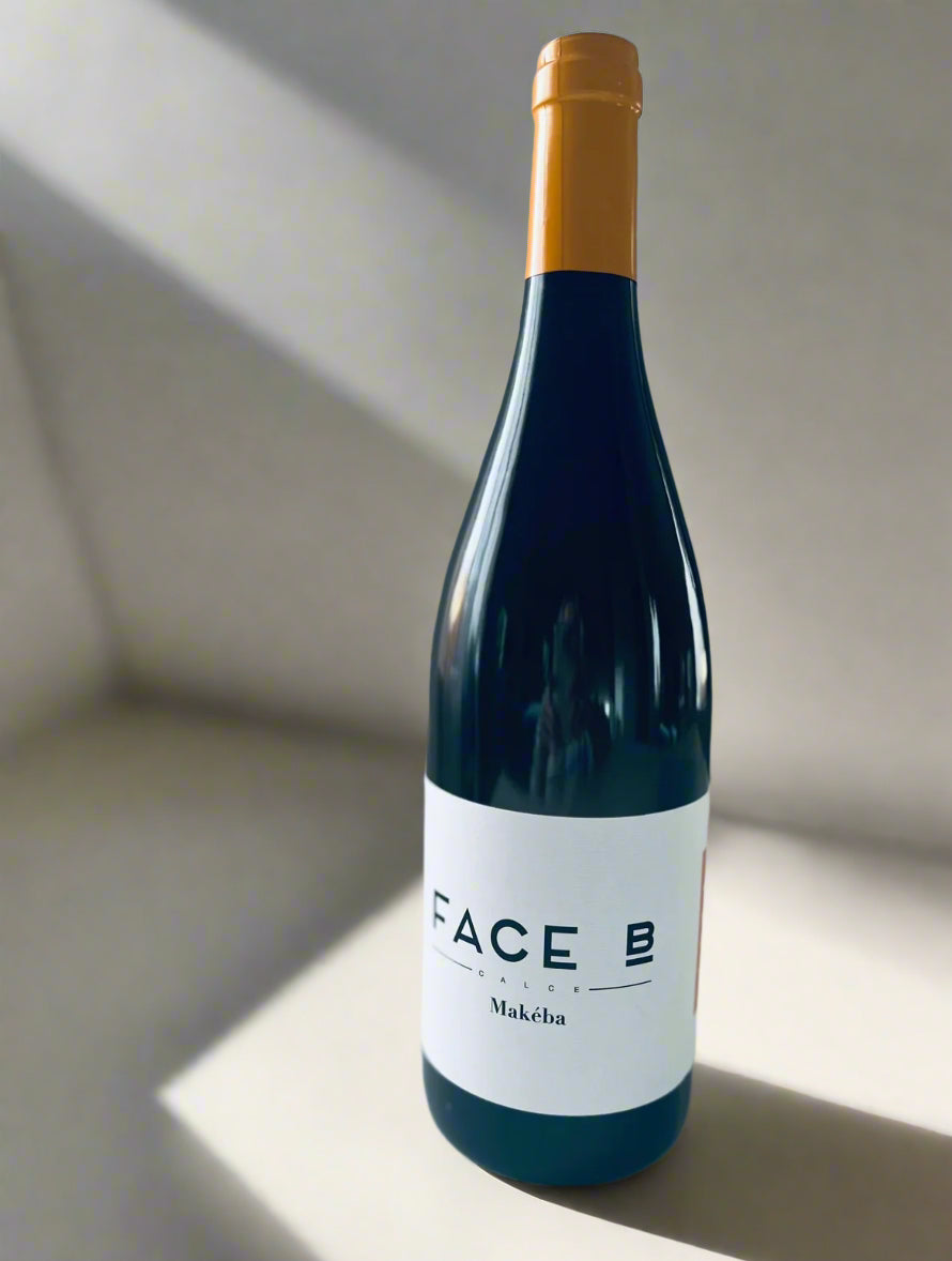 A bottle of Face B Makeba white wine with a minimalist white label, featuring a golden cap, placed on a light surface against a soft grey background.