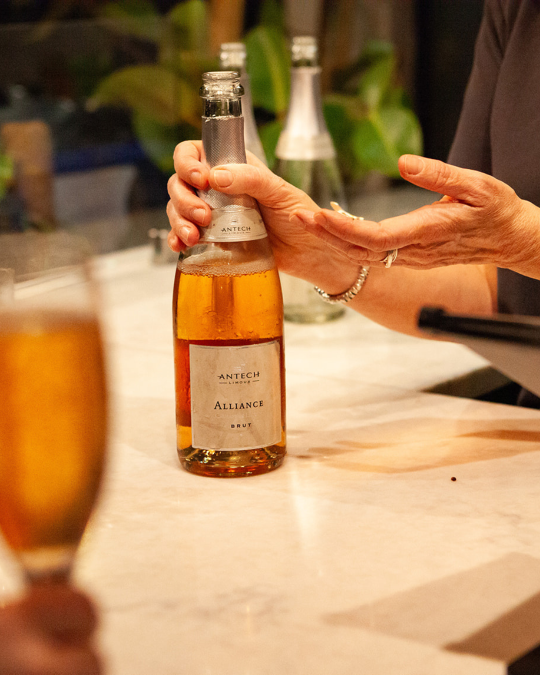 Close-up of a hand presenting a bottle of Antech Alliance Brut sparkling rosé wine on a marble counter, with blurred glass in the foreground and bottles in the background