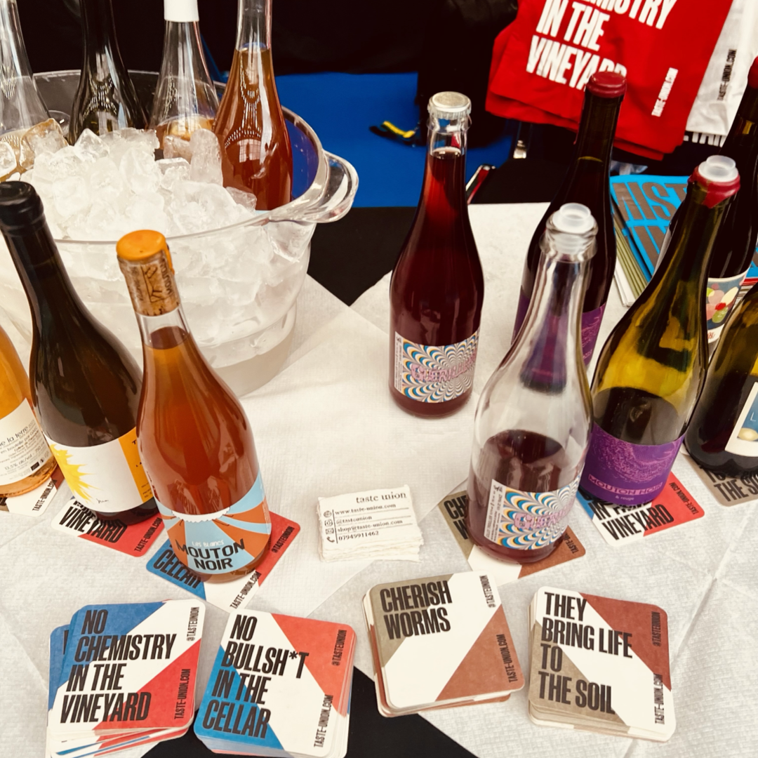 A wine display featuring chilled bottles with colourful labels on a white tablecloth, surrounded by coasters with eco-friendly slogans, and tote bags in the background promoting vineyard culture, emphasising sustainable winemaking practices.