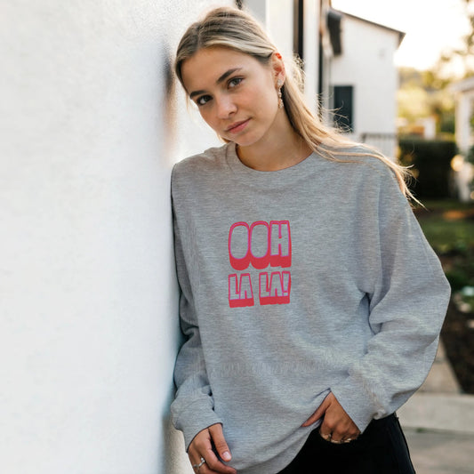 Woman wearing grey Ooh La La sweatshirt by Taste Union, relaxed fit unisex jumper with pink retro lettering, casual outdoor lifestyle photo