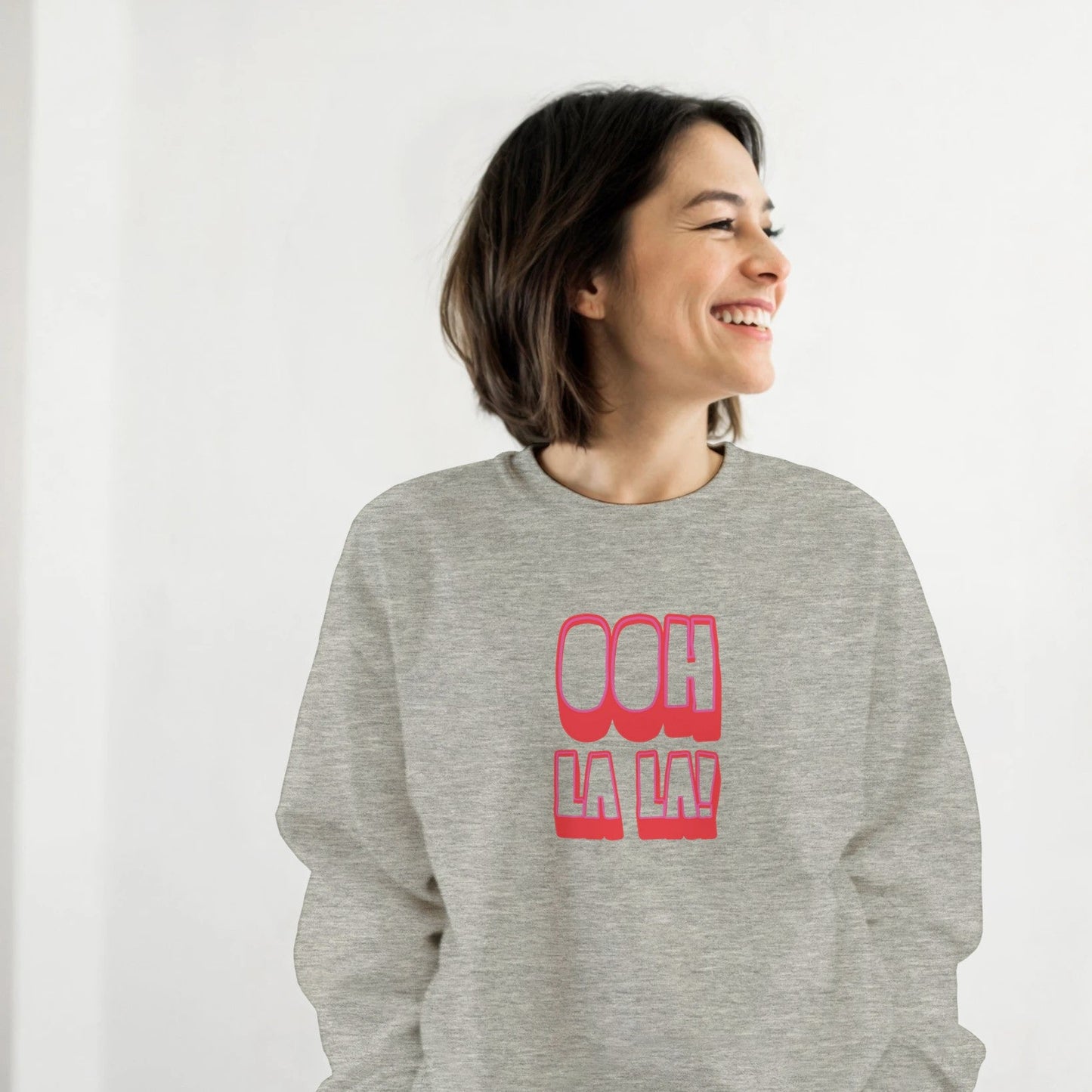 Woman smiling while wearing grey Ooh La La sweatshirt by Taste Union, unisex crewneck jumper with pink retro lettering, relaxed indoor lifestyle photo