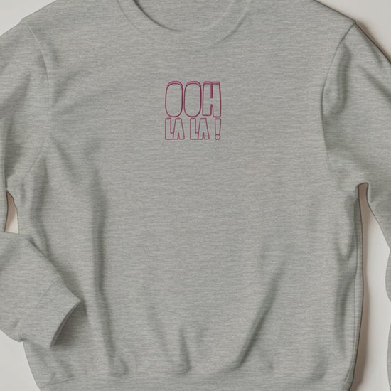 A woman at home wearing the pink “Ooh La La” sweater, smiling as she talks about how fabulous it feels. The cosy setting and natural lighting highlight the sweater’s relaxed, confident vibe — capturing warmth, comfort, and effortless style.