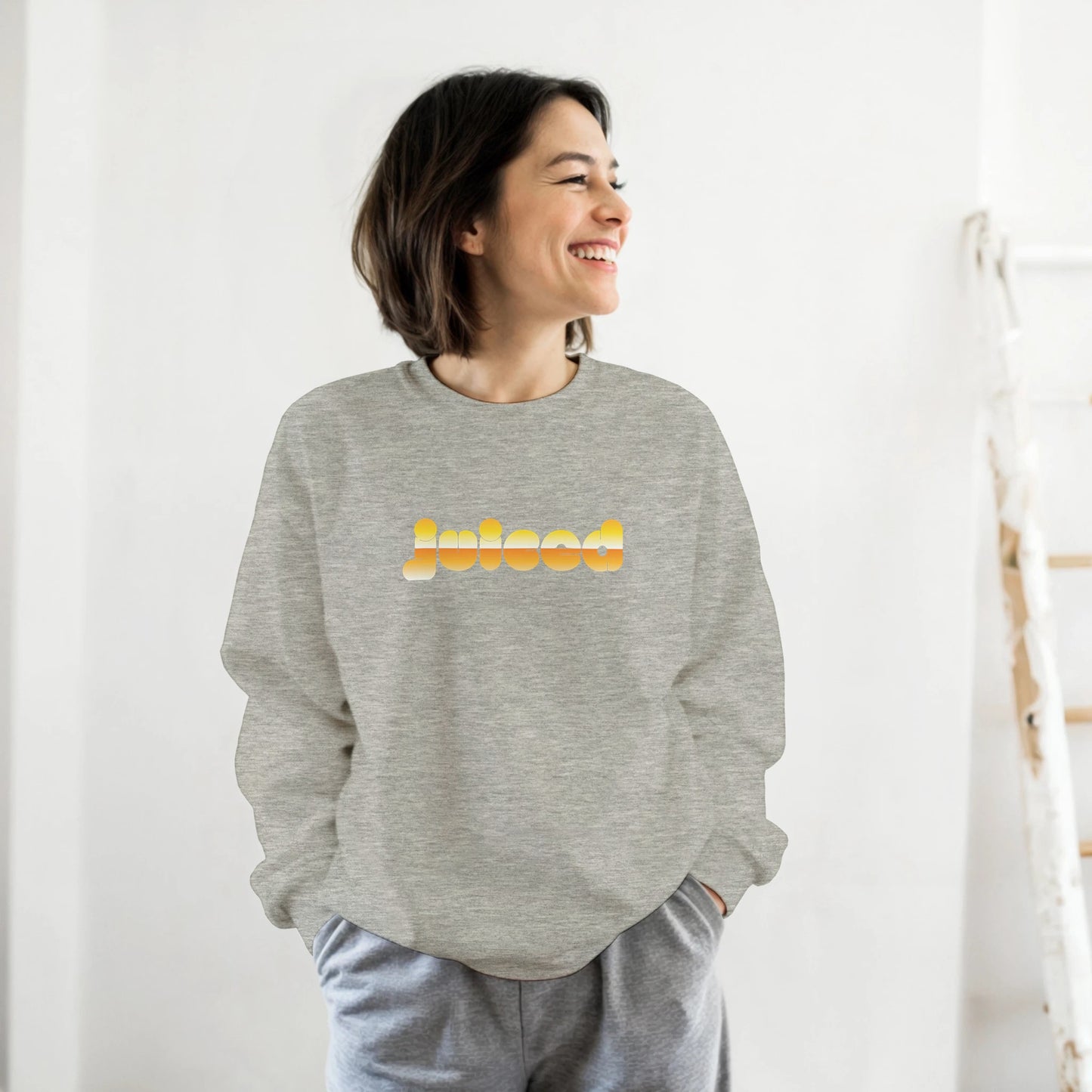Woman wearing the Juiced sweatshirt by Taste Union — unisex grey crewneck with yellow text design inspired by natural wine culture, photographed in a bright modern interior.