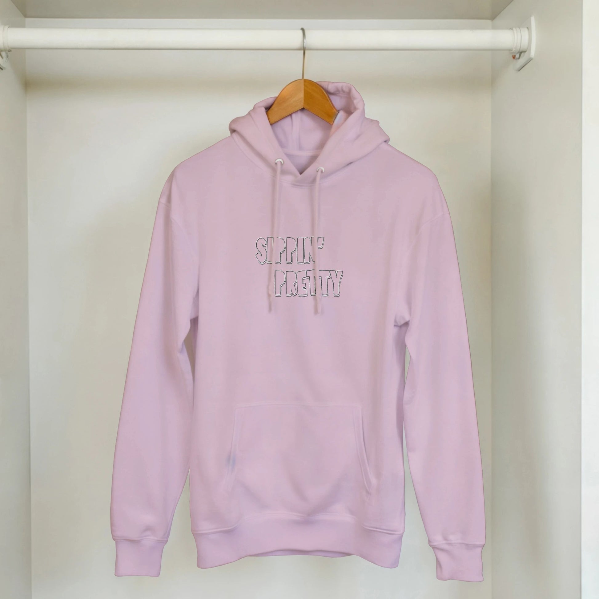 Pink embroidered hoodie with white stitched ‘Sippin’ Pretty’ text hanging on wooden hanger. Sustainable unisex clothing by Taste Union.