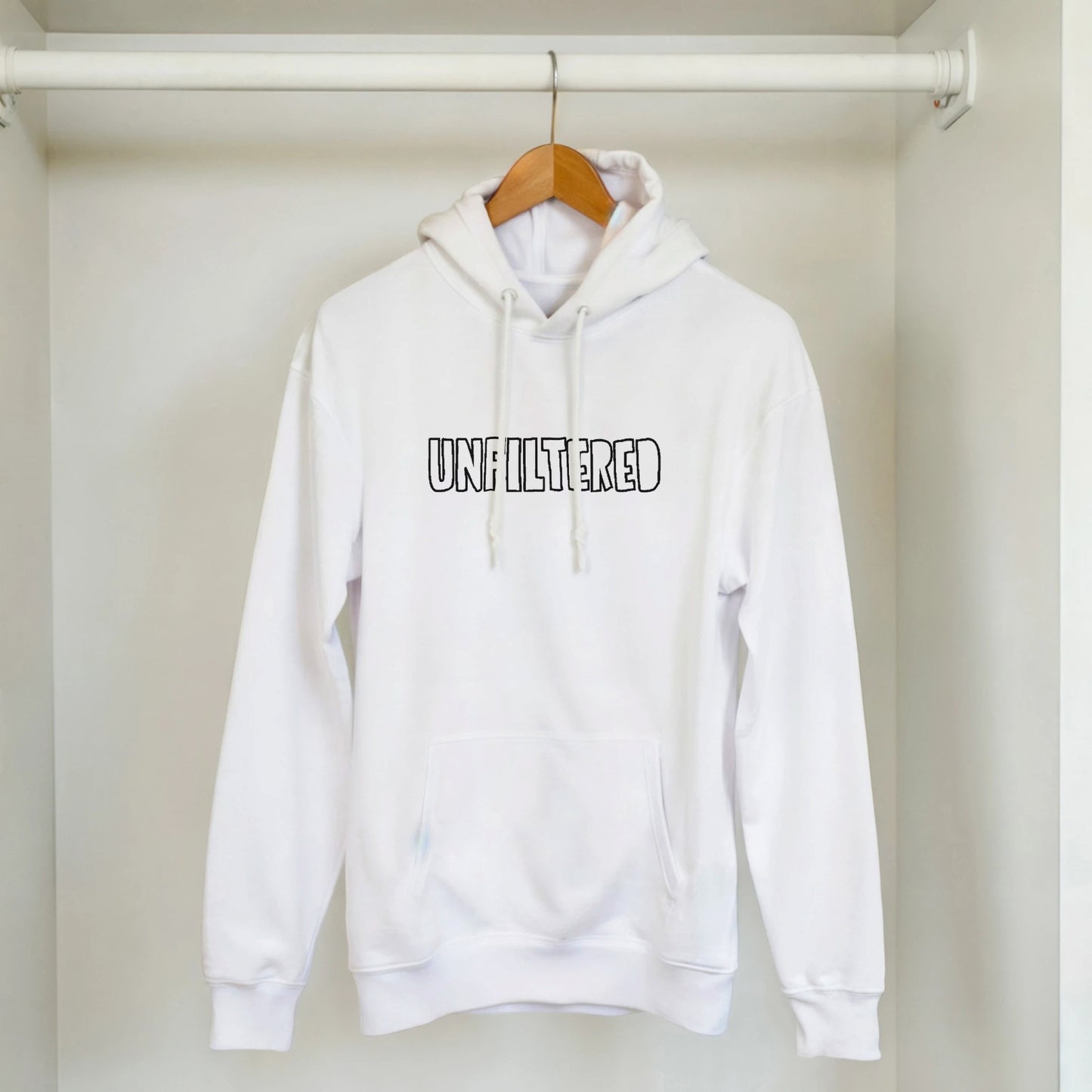White unisex hoodie with black embroidery reading ‘Unfiltered’ hanging on wooden hanger. Minimal sustainable style by Taste Union.