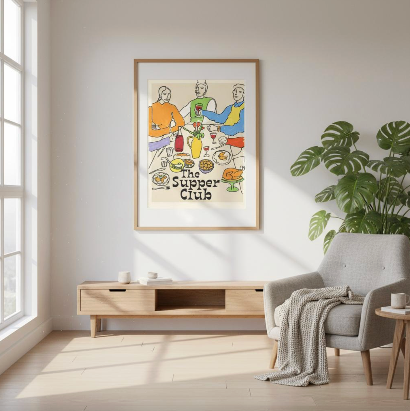 Framed “The Supper Club” art print hanging in a bright modern living room with a wooden console, grey armchair, and large plant. Colourful wine-and-dining illustration adding warmth and personality to contemporary home decor.