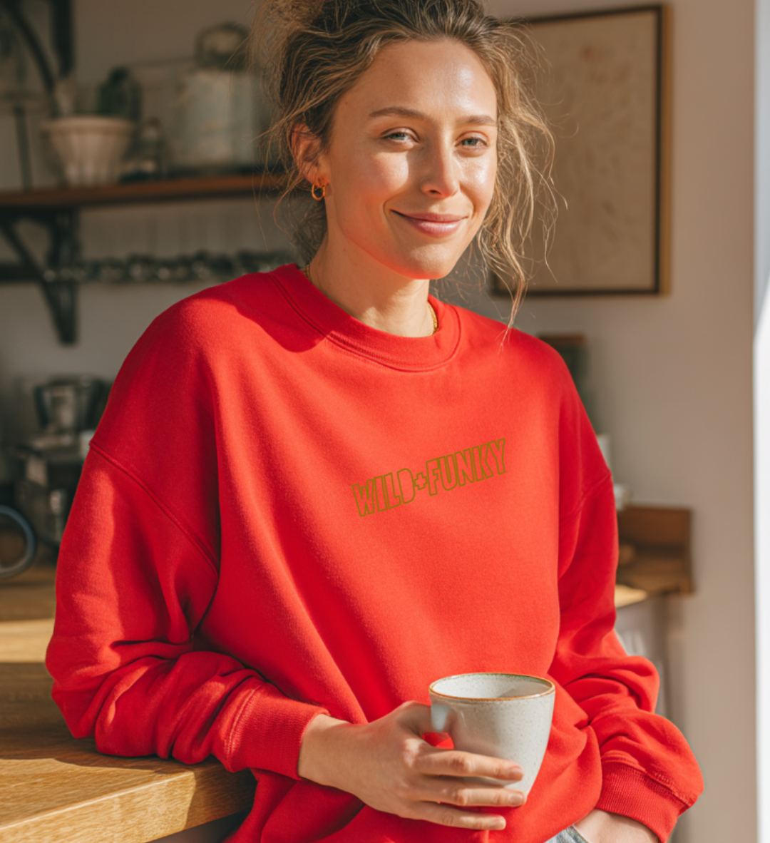 Woman in a bright red ‘Wild + Funky’ embroidered sweatshirt holding a mug in a warm kitchen setting — cosy lifestyle shot for Taste Union apparel.