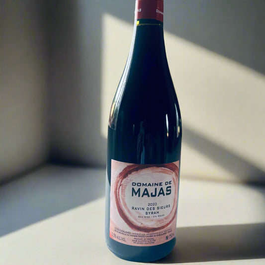 Bottle of Domaine de Majas Ravin des Sieurs 2020 Syrah wine, showcasing a vibrant label on a ruby-red wine, placed on a white table with a soft background.