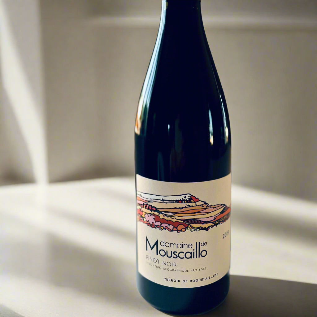 Bottle of Domaine de Mouscaillo Pinot Noir 2019 with label featuring colourful vineyard artwork, placed on a light surface in soft lighting.